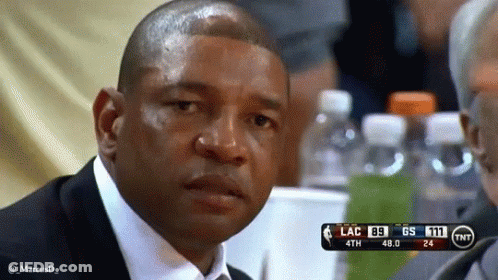 A Man In A Suit And Tie Is Sitting At A Table Watching A Basketball Game With A Scoreboard Behind Him . GIF