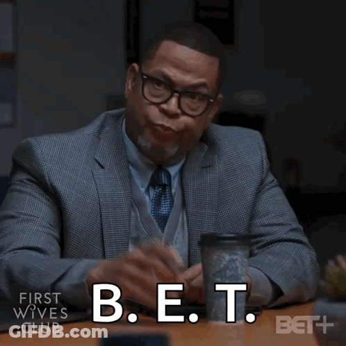 A Man In A Suit And Tie Is Sitting At A Table With A Cup Of Coffee And The Words B.e.t. Below Him GIF