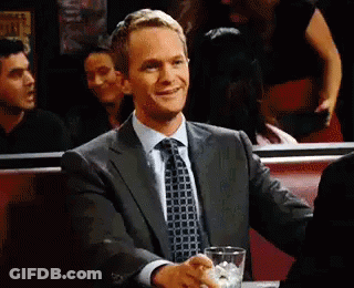 A Man In A Suit And Tie Is Sitting At A Table With A Drink . GIF