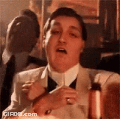 A Man In A Suit And Tie Is Sitting At A Table With A Glass Of Wine In His Hand . GIF