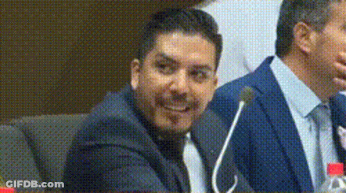 A Man In A Suit And Tie Is Sitting At A Table With A Microphone . GIF