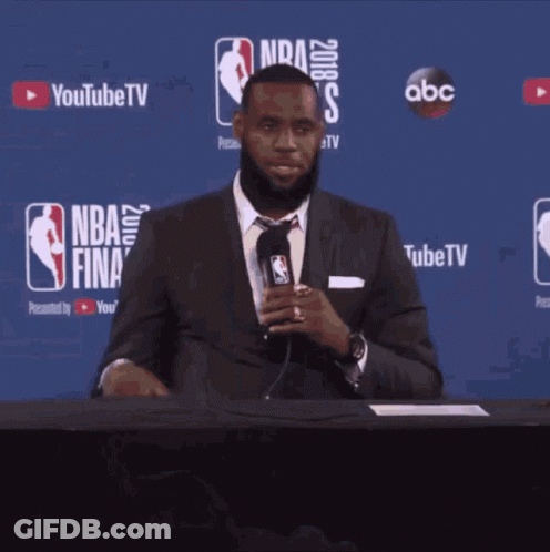 A Man In A Suit And Tie Is Sitting At A Table With A Microphone In Front Of A Nba Finals Sign GIF