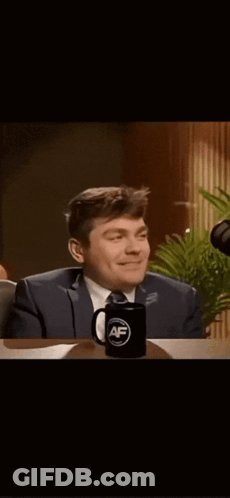 A Man In A Suit And Tie Is Sitting At A Table With A Mug That Says ' Af ' On It GIF