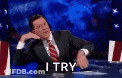 A Man In A Suit And Tie Is Sitting At A Table With A Sign That Says I Try . GIF