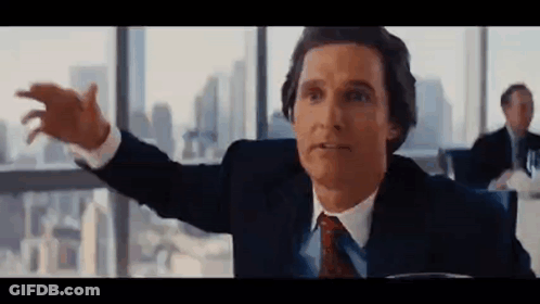 A Man In A Suit And Tie Is Sitting At A Table With His Arms Outstretched In Front Of A Window . GIF