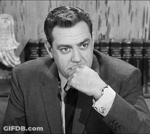 A Man In A Suit And Tie Is Sitting At A Table With His Hand On His Chin And Looking At The Camera . GIF