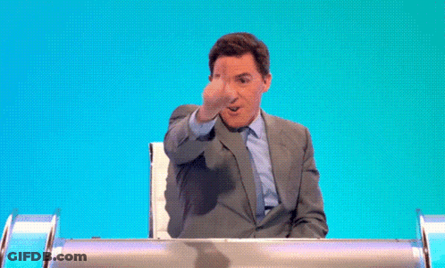 A Man In A Suit And Tie Is Sitting At A Table With His Hand Up . GIF
