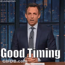 A Man In A Suit And Tie Is Sitting At A Table With His Hands Folded And Says Good Timing . GIF