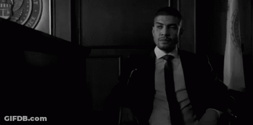 A Man In A Suit And Tie Is Sitting At A Table With His Hands Folded In A Courtroom . GIF