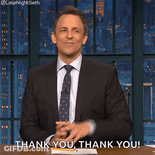 A Man In A Suit And Tie Is Sitting At A Table With His Hands In The Air And Saying Thank You . GIF