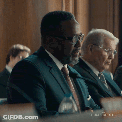 A Man In A Suit And Tie Is Sitting At A Table With Thunderbolts Written On The Bottom Right GIF
