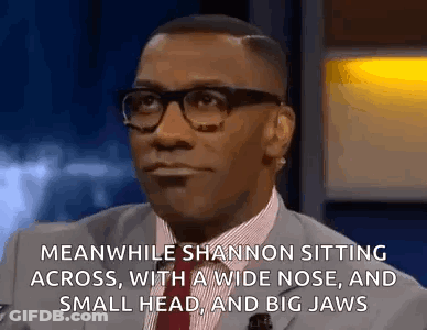 A Man In A Suit And Tie Is Sitting Down With A Wide Nose And A Small Head And Big Jaws . GIF