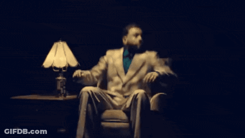 A Man In A Suit And Tie Is Sitting In A Chair In Front Of A Lamp GIF