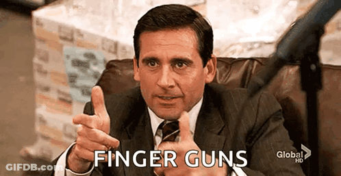 A Man In A Suit And Tie Is Sitting In A Chair Pointing At The Camera And Saying Finger Guns . GIF