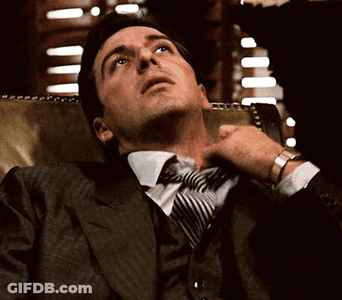 A Man In A Suit And Tie Is Sitting In A Chair GIF