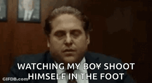 A Man In A Suit And Tie Is Sitting In A Chair Watching His Boy Shoot Himself In The Foot . GIF