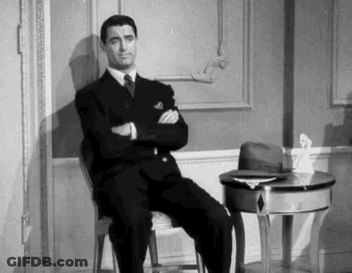 A Man In A Suit And Tie Is Sitting In A Chair With His Arms Crossed In A Room . GIF
