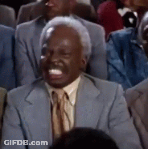 A Man In A Suit And Tie Is Sitting In A Crowd Of People And Laughing . GIF
