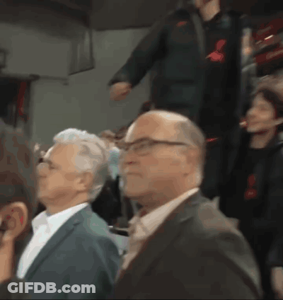 A Man In A Suit And Tie Is Sitting In A Crowd Of People GIF