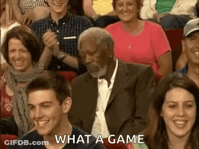 A Man In A Suit And Tie Is Sitting In A Crowd Of People Laughing . GIF