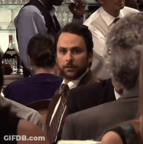 A Man In A Suit And Tie Is Sitting In A Crowded Room GIF