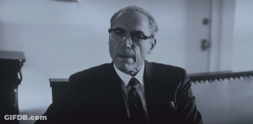 A Man In A Suit And Tie Is Sitting In A Room GIF