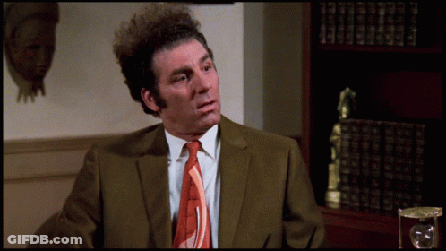A Man In A Suit And Tie Is Sitting In Front Of A Bookcase GIF