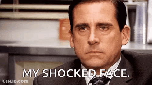 A Man In A Suit And Tie Is Sitting In Front Of A Counter And Says `` My Shocked Face '' . GIF