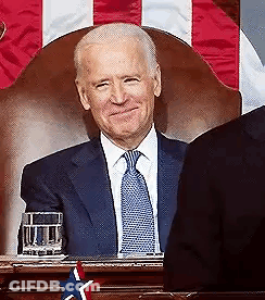A Man In A Suit And Tie Is Sitting In Front Of A Flag . GIF