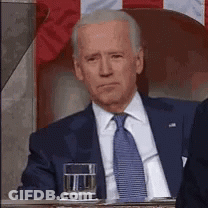 A Man In A Suit And Tie Is Sitting In Front Of A Flag With A Glass Of Water . GIF