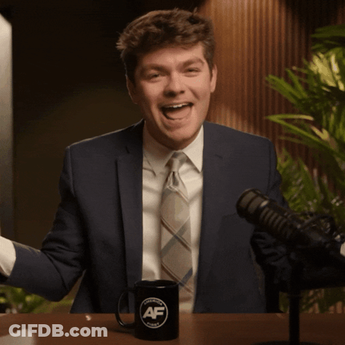 A Man In A Suit And Tie Is Sitting In Front Of A Microphone And A Mug That Says Af GIF