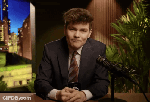 A Man In A Suit And Tie Is Sitting In Front Of A Microphone With A Cell Phone In His Hand GIF