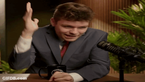 A Man In A Suit And Tie Is Sitting In Front Of A Microphone With A Coffee Mug On The Table GIF