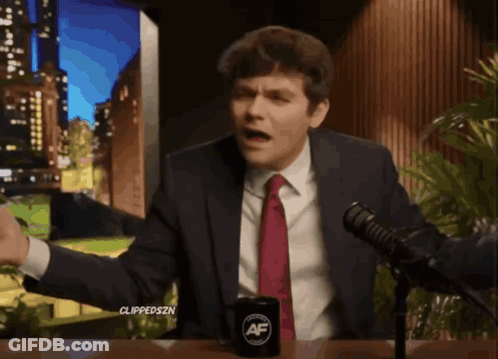 A Man In A Suit And Tie Is Sitting In Front Of A Microphone With A Cup That Says ' Af ' On It GIF