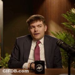 A Man In A Suit And Tie Is Sitting In Front Of A Microphone With A Mug That Says Af On It GIF