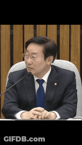 A Man In A Suit And Tie Is Sitting In Front Of A Microphone With His Hands Folded GIF