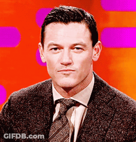 A Man In A Suit And Tie Is Sitting In Front Of A Purple Background GIF