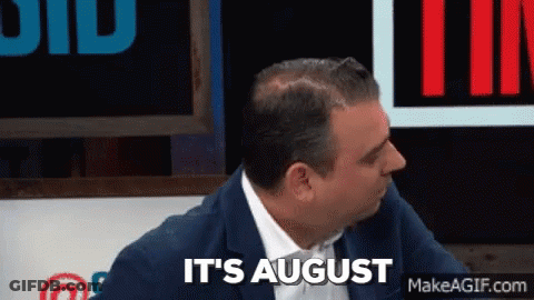 A Man In A Suit And Tie Is Sitting In Front Of A Sign That Says It 'S August GIF