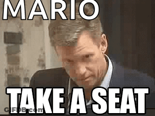 A Man In A Suit And Tie Is Sitting In Front Of A Sign That Says Mario Take A Seat . GIF