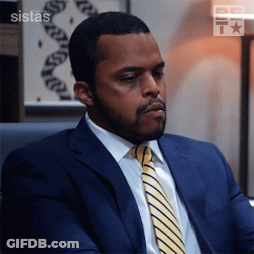 A Man In A Suit And Tie Is Sitting In Front Of A Sign That Says ' Sistas ' GIF
