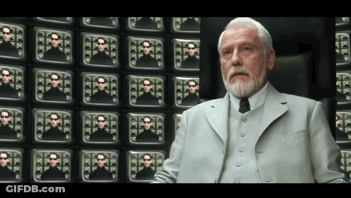 A Man In A Suit And Tie Is Sitting In Front Of A Wall Of Television Screens GIF