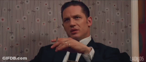 A Man In A Suit And Tie Is Sitting In Front Of A Wall With A Floral Pattern GIF