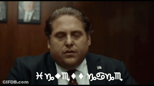 A Man In A Suit And Tie Is Sitting In Front Of A Wall With A Sign That Says Hnm On It GIF