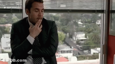 A Man In A Suit And Tie Is Sitting In Front Of A Window And Covering His Mouth With His Hand . GIF