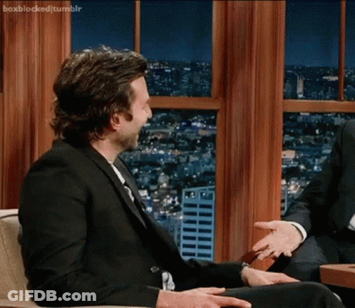 A Man In A Suit And Tie Is Sitting In Front Of A Window Talking To Another Man .. GIF