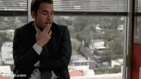 A Man In A Suit And Tie Is Sitting In Front Of A Window With His Hand On His Chin . GIF