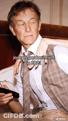 A Man In A Suit And Tie Is Sitting On A Bed With A Woman Holding His Arm . GIF