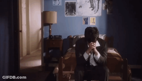 A Man In A Suit And Tie Is Sitting On A Bed With His Hands Folded . GIF