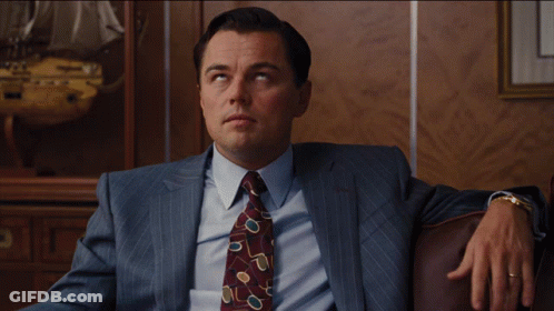 A Man In A Suit And Tie Is Sitting On A Brown Couch GIF