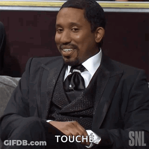A Man In A Suit And Tie Is Sitting On A Couch And Touching His Chest With The Snl Logo Behind Him GIF